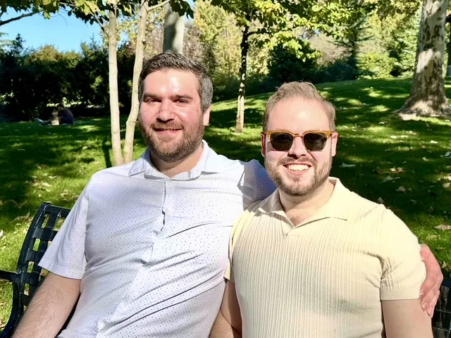 Zack and Mason sitting close on a park bench under sunlit trees.